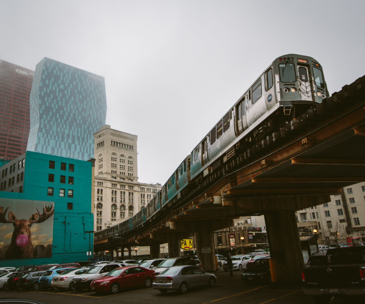 An Orange Line passenger train to Midway rolls through the elevated s-curved track just south of the loop. One of many unique billboards in Chicago over looks the parking lot from the side of a turquoise building of a moose blowing a pink bubble gum bubble.